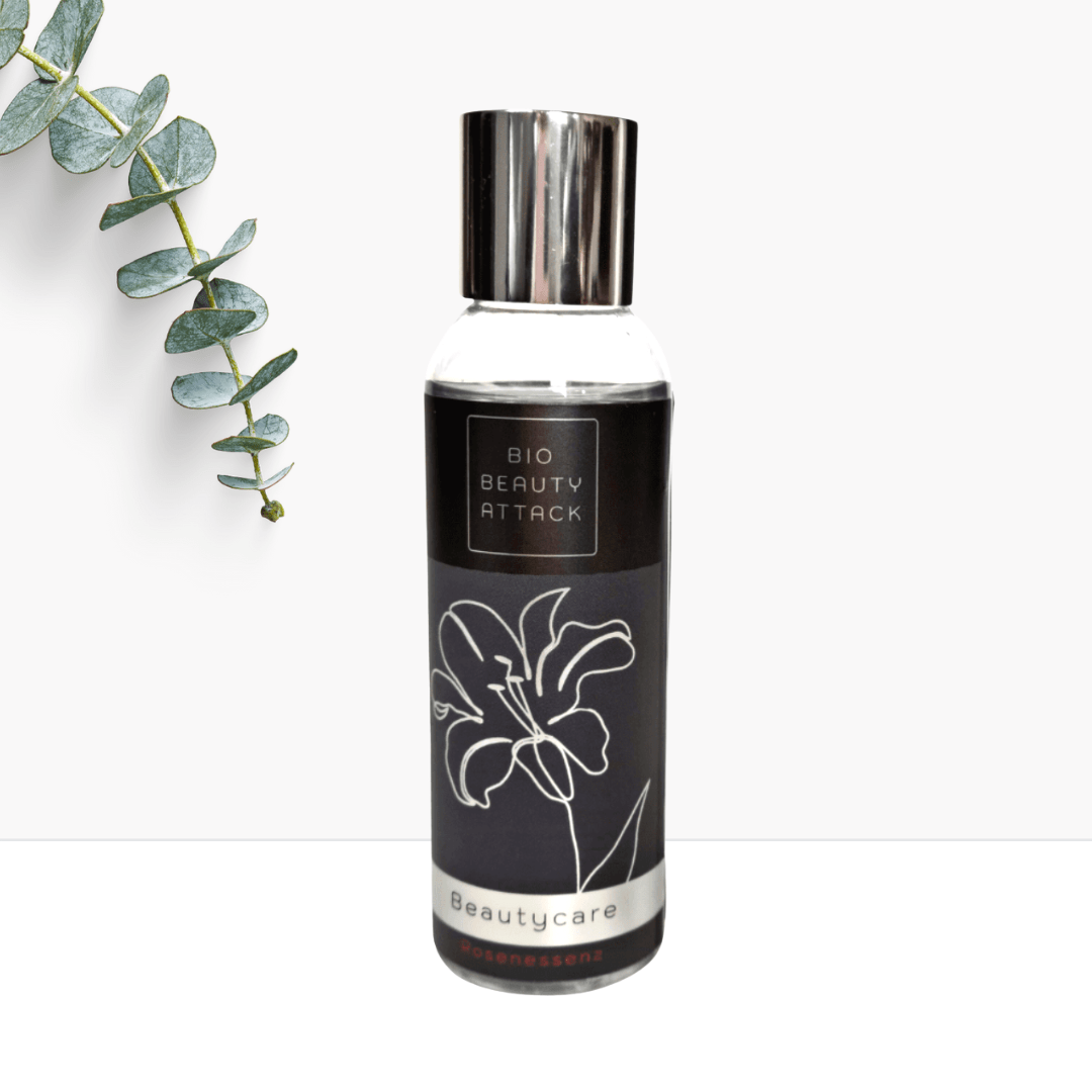 Flasche Bio Beauty Attack Rosenessenz mit schwarzem Etikett, silbernem Deckel und Blütenmotiv, dekoriert mit Eukalyptuszweig auf weißem Hintergrund.