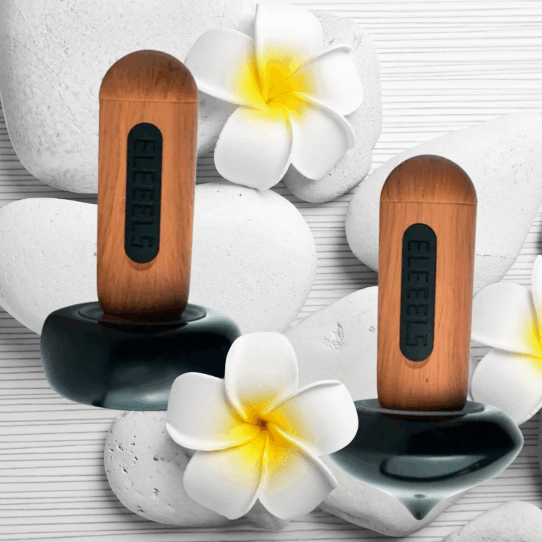 Zwei ELEEELS Wärme-Stäbe liegen auf weißen Steinen mit Blüten. Moderne Hot-Stone-Technologie für Massage, Wellness und Tiefenentspannung in Gütersloh.