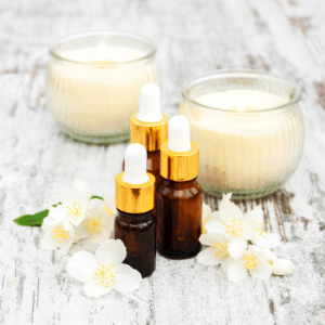 Drei kleine braune Glasfläschchen mit goldenen Pipetten stehen auf einem hellen Holztisch, umgeben von weißen Blüten und zwei brennenden Kerzen im Glas. Das Bild vermittelt Ruhe, Entspannung und natürliche Pflege.