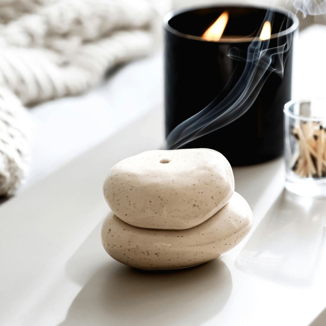 Zwei helle, glatte Steinobjekte liegen übereinander und dienen als schlichte Räucherschale. Aus der oberen Schale steigt zarter Rauch auf. Im Hintergrund brennt eine schwarze Duftkerze und sorgt für eine warme, entspannte Atmosphäre.