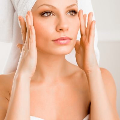 Eine Frau mit Handtuch im Haar berührt sanft ihre Schläfen und schaut entspannt in den Spiegel. Das Bild steht für eine wohltuende Access Facelift Behandlung in Gütersloh, die natürliche Schönheit und innere Ruhe fördert.