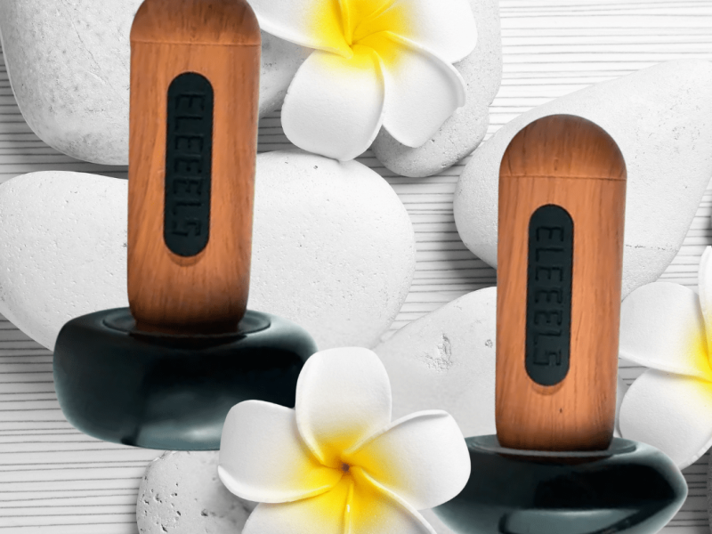 Zwei ELEEELS Wärme-Stäbe liegen auf weißen Steinen mit Blüten. Moderne Hot-Stone-Technologie für Massage, Wellness und Tiefenentspannung in Gütersloh.